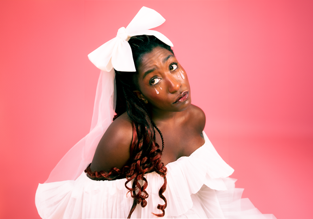Sophie Duker, a young black woman, is in front of a baby pink background. Her hair is styled in braids, and curled on the bottom, she is wearing a white bow keeping her braids away from her face. She has pink tears on both cheeks.