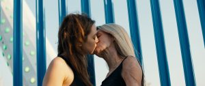 White and blue striped background. Two white females kissing. One with long brown hair wearing a black vest to the left and one with long blonde hair, visible burn scars wearing a black vest.