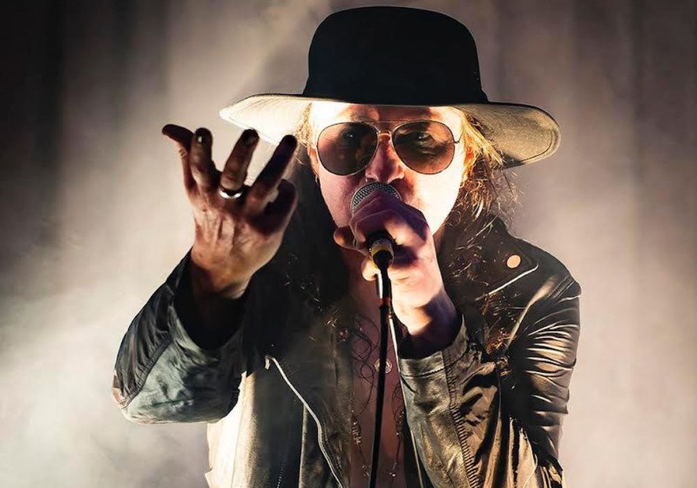 Image of Dave from Sanctum Sanctorium, he wears a wide brimmed hat and leather coat and holds hi hand out as if holding something unseen with one hand and holding a microphone to his mouth in the other.
