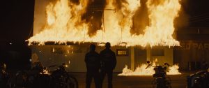 Two male figures dressed in bikers leathers looking on as a building is on fire.