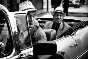 Black and White image of two males sat in an open-topped car. One in the front, one in the rear. Both are wearing suits and Panama hats.