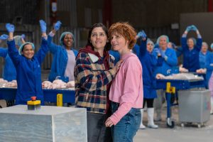 Workers in a chicken factory dressed in blue uniforms and blue hair nets. There are two females in the centre stood close to each other. To the right is white with messy bobbed auburn hair wearing a pink jumper and blue jeans. To the left white with dark hair wearing a checked coat.