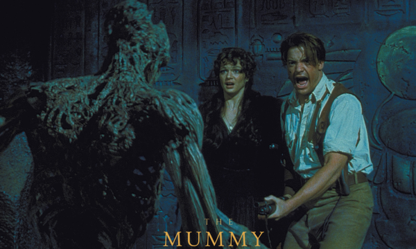 White male with mousey hair wearing a white shirt and cream trousers and waistcoat. A white female with long dark hair wearing black. The main is holding a gun at a mummy/ zombie.
