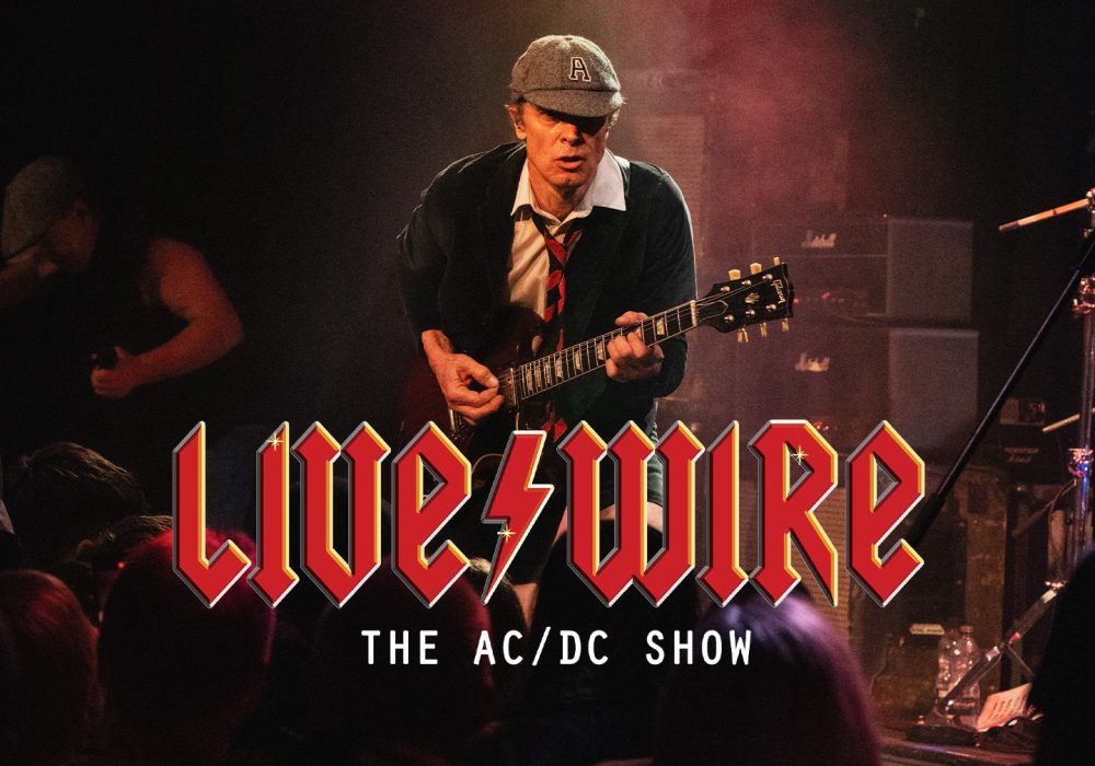 A guitarist performing on stage, dressed in a school uniform-inspired outfit with a cap, white shirt, dark sweater, and red tie. He's playing a Gibson SG guitar, leaning forward in an energetic pose. The background shows Marshall amplifiers and moody stage lighting. Below the image is the text 'Live/Wire: The AC/DC Show' in a red, angular font reminiscent of AC/DC's iconic logo.