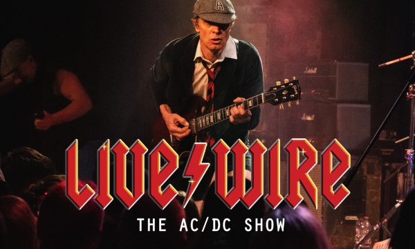 A guitarist performing on stage, dressed in a school uniform-inspired outfit with a cap, white shirt, dark sweater, and red tie. He's playing a Gibson SG guitar, leaning forward in an energetic pose. The background shows Marshall amplifiers and moody stage lighting. Below the image is the text 'Live/Wire: The AC/DC Show' in a red, angular font reminiscent of AC/DC's iconic logo.