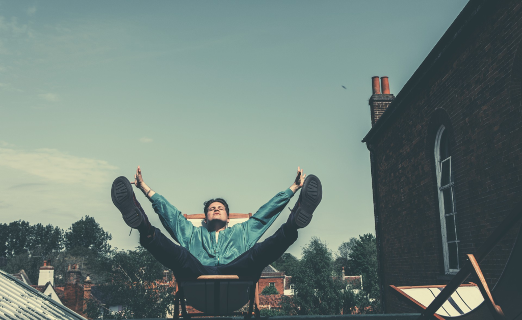 Luke, sitting in a deckchair on a rooftop terrace, with his eyes closed and his arms and legs raised.