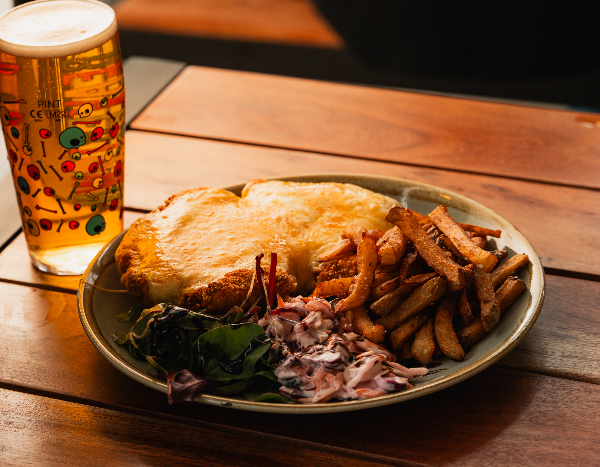 Image of a chicken parmo and chips on a green plate. The plate is sat on a slatted wooden surface with a pint of amber beer in a glass with colourful skulls across it.