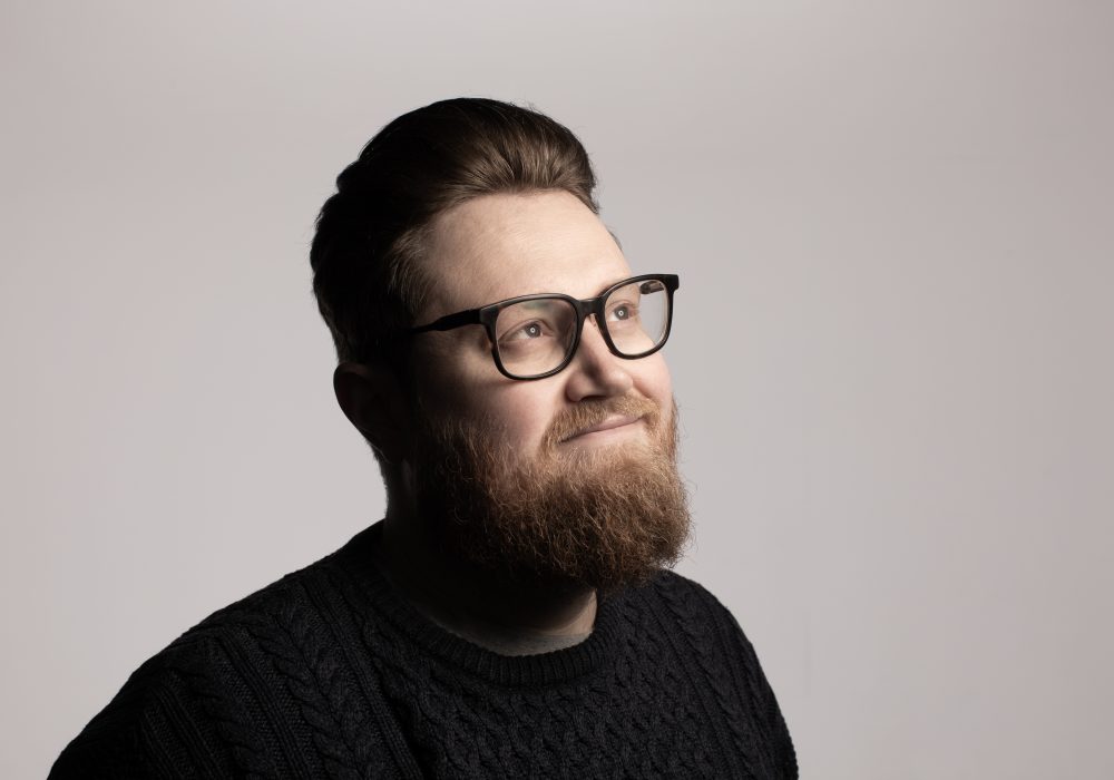 A headshot of comedian Pete Selwood, a white man with short brown hair, black rimmed glasses, and a brown beard. He is looking off to the right, and is smiling.