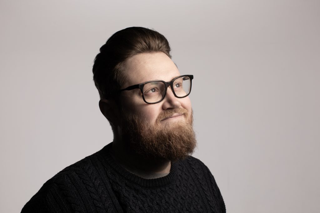 A headshot of comedian Pete Selwood, a white man with short brown hair, black rimmed glasses, and a brown beard. He is looking off to the right, and is smiling.