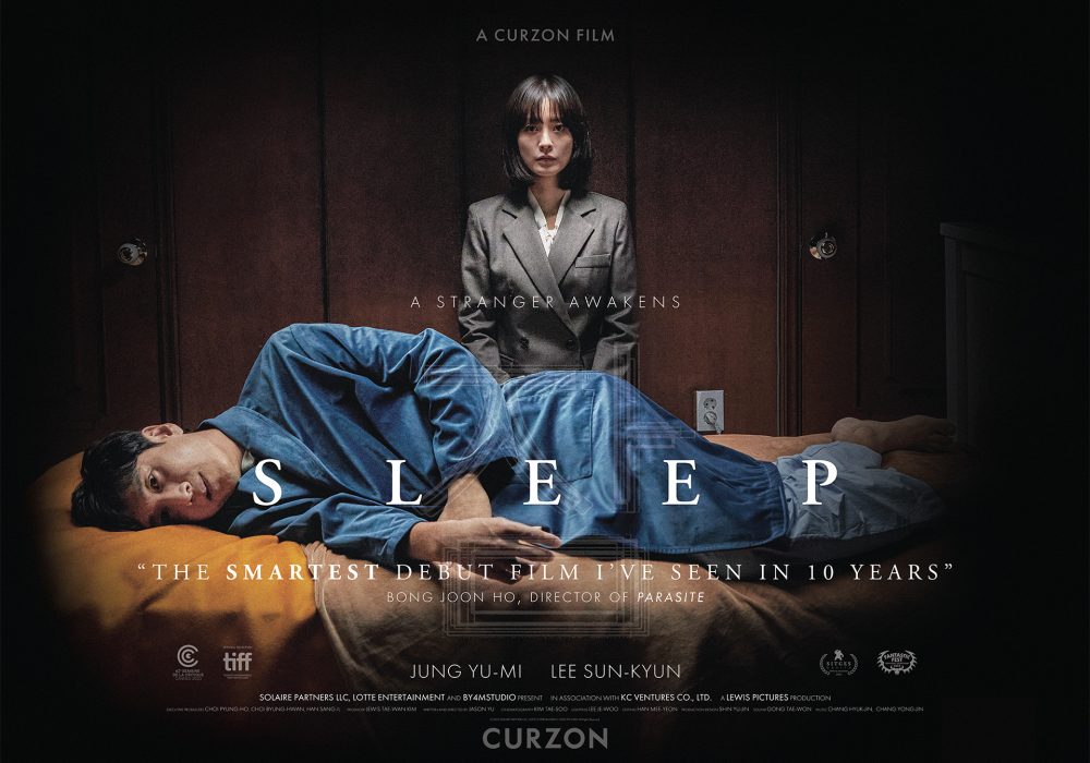 An Asian man with black hair laying side ways on a bed wearing blue pyjamas and a blue dressing gown with an Asian woman with a black bob and fringe wearing a grey suit stood behind him.