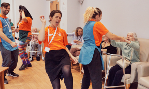 Image shows the cast of The Home Roadshow in a care home mid performance, the cast wear bright orange and turquoise clothing and are dancing and interacting with the care home residents.