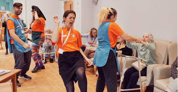 Image shows the cast of The Home Roadshow in a care home mid performance, the cast wear bright orange and turquoise clothing and are dancing and interacting with the care home residents.