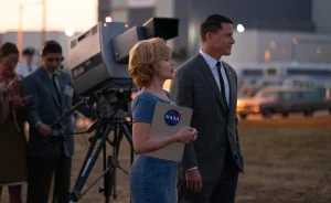 Image of Scarlett Johansson a white female with blonde hair wearing a blue dress holding a NASA clipboard and Channing Tatum a white male with dark hair wearing a grey suit, white shirt and dark tie. 