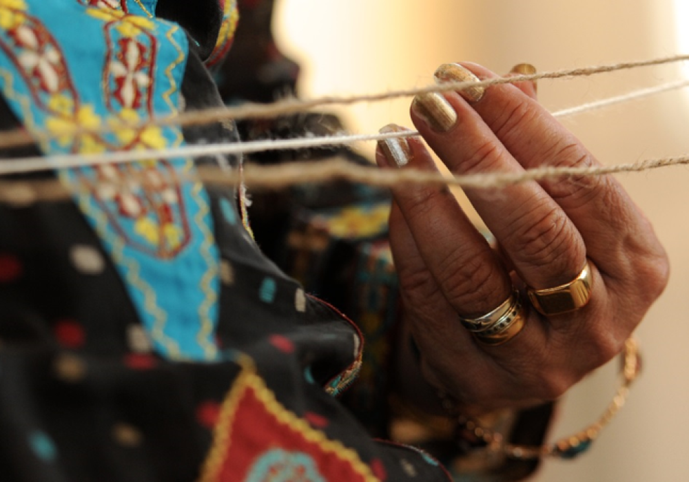 Close up of a hand, with gold rings, holding one of three pieces of string which appear horizontally across the width of the picture