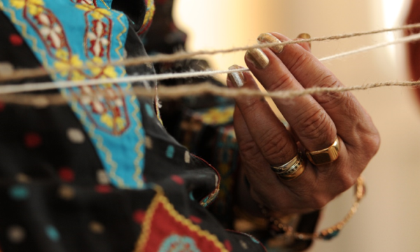 Close up of a hand, with gold rings, holding one of three pieces of string which appear horizontally across the width of the picture