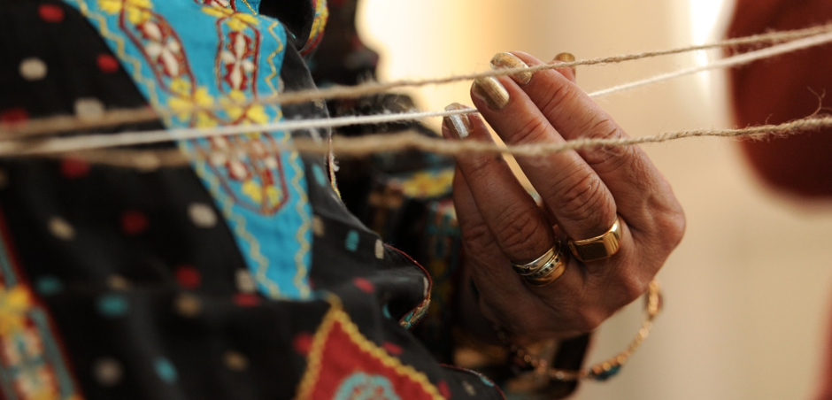 Close up of a hand, with gold rings, holding one of three pieces of string which appear horizontally across the width of the picture