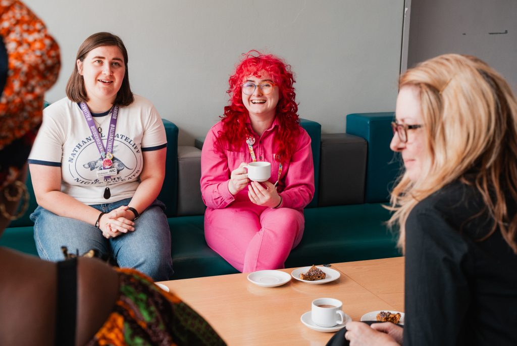 Several people sitting around a table, chatting, drinking coffee, and eating cake. The photo focuses on Allison, one of ARC's Producers. She is wearing a bright pink jumpsuit and has bright pink hair.