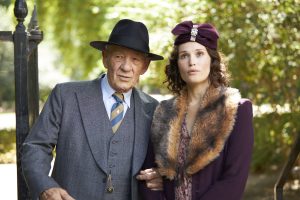 Ian McKellen wearing a blue suit with a blue bowler hate linked arm in arm with Gemma Arterton a white female with bob length brown hair wearing a burgandy hat with a burgundy coat with a big fur collar.