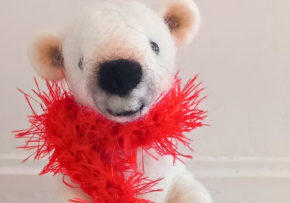 A close-up of a needle felt polar bear, he has light pink ears and nose, and is wearing a bright red fuzzy scarf.