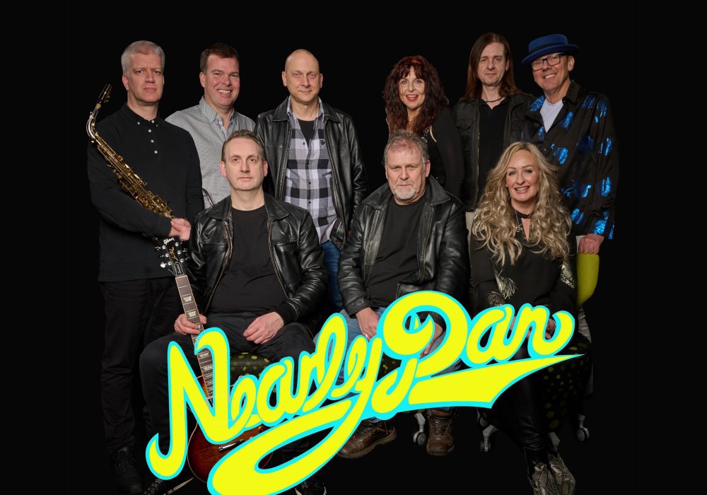 The image shows a group portrait of a band called "Nearly Dan." The band members include a saxophonist, a guitarist, a keyboardist, a female vocalist, and several other musicians. They are posed in front of a large "Nearly Dan" logo in vibrant yellow and blue colors. The members are dressed in a mix of casual and more formal attire, suggesting a professional music group. The image has a dark background, drawing focus to the band members and the logo.