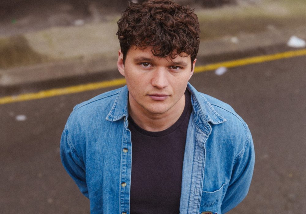 a young man with curly dark hair looking directly at the camera. He has a serious expression on his face. The man is wearing a light blue denim shirt over a dark-colored t-shirt. The background appears to be an outdoor urban setting, with some concrete or pavement visible. The lighting suggests it may be an overcast day. The man's gaze is intense and focused on the viewer.