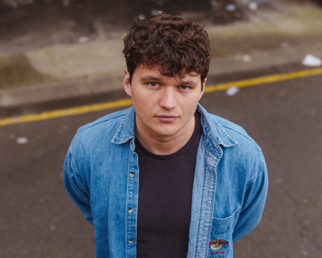 a young man with curly dark hair looking directly at the camera. He has a serious expression on his face. The man is wearing a light blue denim shirt over a dark-colored t-shirt. The background appears to be an outdoor urban setting, with some concrete or pavement visible. The lighting suggests it may be an overcast day. The man's gaze is intense and focused on the viewer.