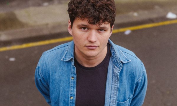 a young man with curly dark hair looking directly at the camera. He has a serious expression on his face. The man is wearing a light blue denim shirt over a dark-colored t-shirt. The background appears to be an outdoor urban setting, with some concrete or pavement visible. The lighting suggests it may be an overcast day. The man's gaze is intense and focused on the viewer.