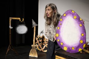 Performer on stage holding a large cut out egg. A broomstick with a witch's hat on top of it is off to the side.