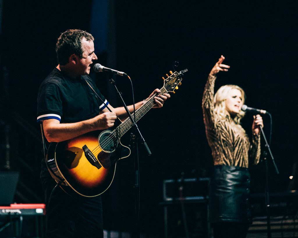 a live music performance on stage. In the foreground, there's a man playing an acoustic guitar and singing into a microphone. He's wearing a dark shirt with light-colored stripes on the sleeves. To his right, there's a woman with long blonde hair singing into another microphone, with her arm raised expressively. In the background on the left, there's a person seated at a keyboard, which appears to be a Nord brand synthesizer based on its red color. The lighting creates a dramatic atmosphere, with the performers illuminated against a dark background. The guitarist's instrument appears to be a sunburst-colored acoustic-electric guitar. The overall scene captures the energy and collaboration of a live band performance, with multiple musicians contributing to the sound.