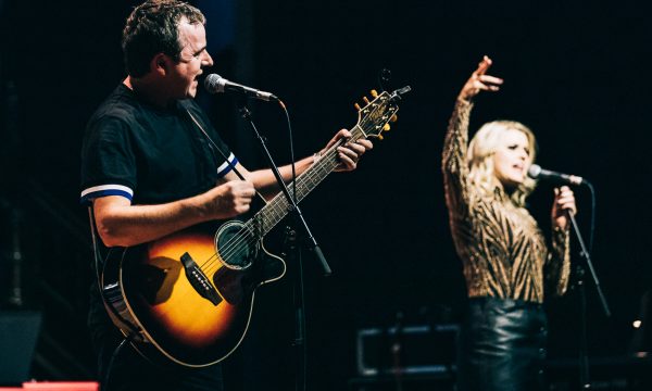 a live music performance on stage. In the foreground, there's a man playing an acoustic guitar and singing into a microphone. He's wearing a dark shirt with light-colored stripes on the sleeves. To his right, there's a woman with long blonde hair singing into another microphone, with her arm raised expressively. In the background on the left, there's a person seated at a keyboard, which appears to be a Nord brand synthesizer based on its red color. The lighting creates a dramatic atmosphere, with the performers illuminated against a dark background. The guitarist's instrument appears to be a sunburst-colored acoustic-electric guitar. The overall scene captures the energy and collaboration of a live band performance, with multiple musicians contributing to the sound.