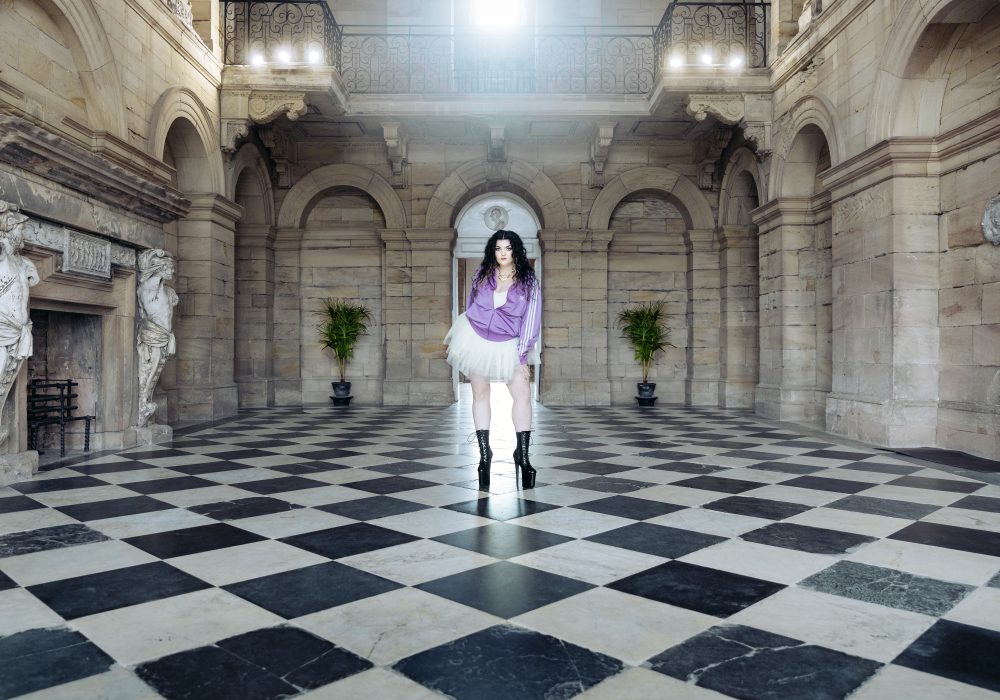 Image of a woman in a tracksuit, tutu, and black boots stood inside a grand stone building.