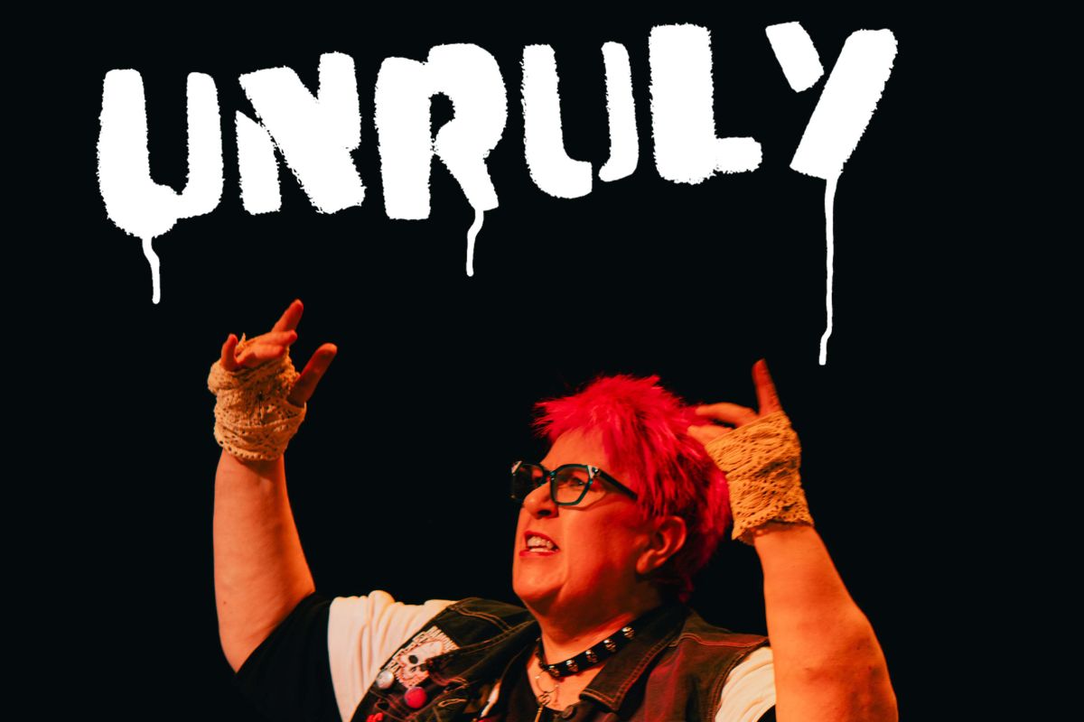 Actress Vici Wreford-Sinnot, with short, bright pink hair, raises her arms up, she has a defiant expression. Above her is the word Unruly, in white spray paint.