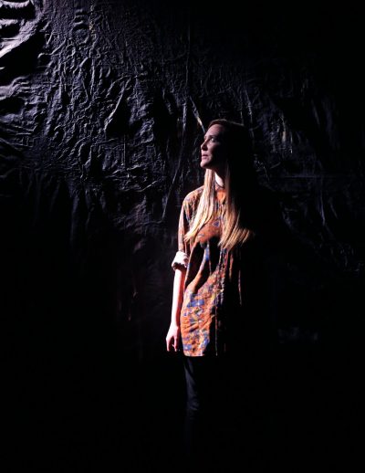 ARC Associate Artist stands in partial darkness, against a black wall. She is looking up to her right, from where a light source casts light on her face and the upper part of her body.